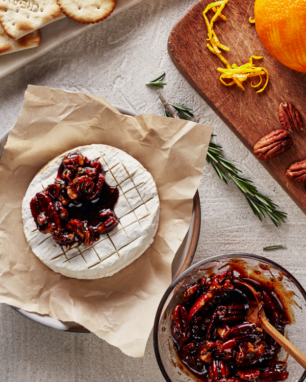 Brie cuit au four, garni de noix de pécan confites épicées et de sirop de Cassonade Foncée, décoré de zeste d'orange et de romarin, présenté dans une assiette sur un plateau de service avec des craquelins, des crostinis et des tranches de poires et de pommes, avec des verres à vin en arrière-plan.