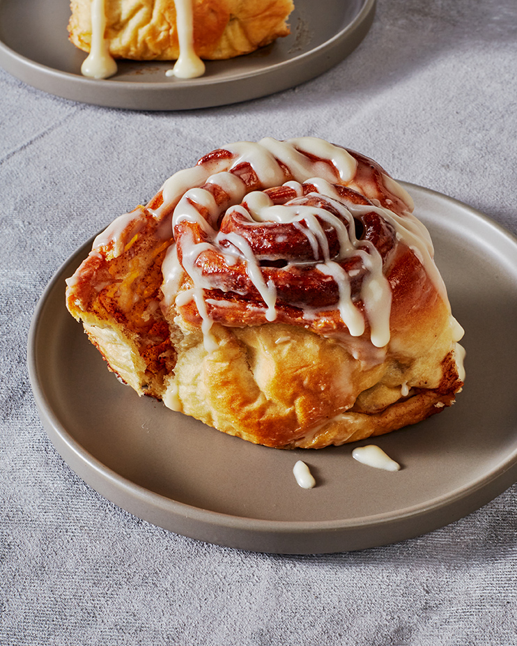 Un petit pain à la cannelle arrosé de glaçage sur une assiette brune, avec un deuxième petit pain sur une assiette en arrière-plan.