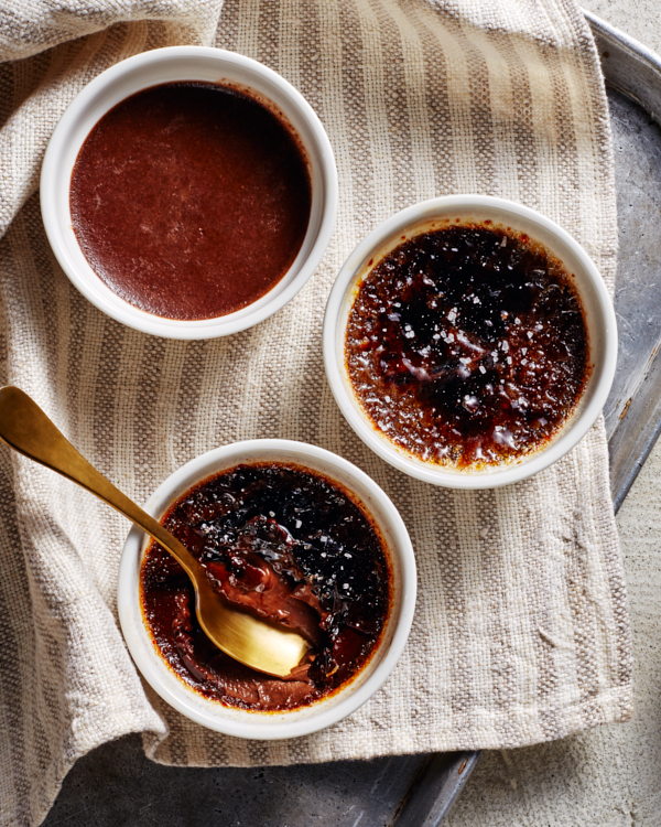 Trois ramequins de crème brûlée au chocolat noir salé, l'un partiellement mangé, l'autre sans garniture, représentés sur un torchon sur une plaque de cuisson avec une cuillère dorée et un bol de Cassonade Dorée.