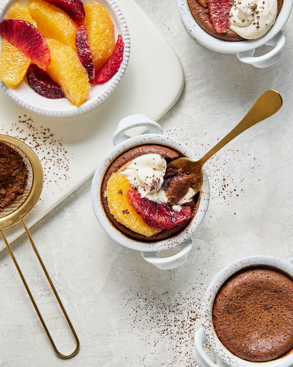  Trois petits gâteaux au chocolat fondant individuels dans des ramequins blancs, dont deux garnis de crème fouettée et de cacao en poudre, agrémentés de segments d'orange et d'orange sanguine, et un nature. Ils sont présentés avec un bol de segments d'orange et d'orange sanguine ainsi qu'un tamis rempli de cacao en poudre.