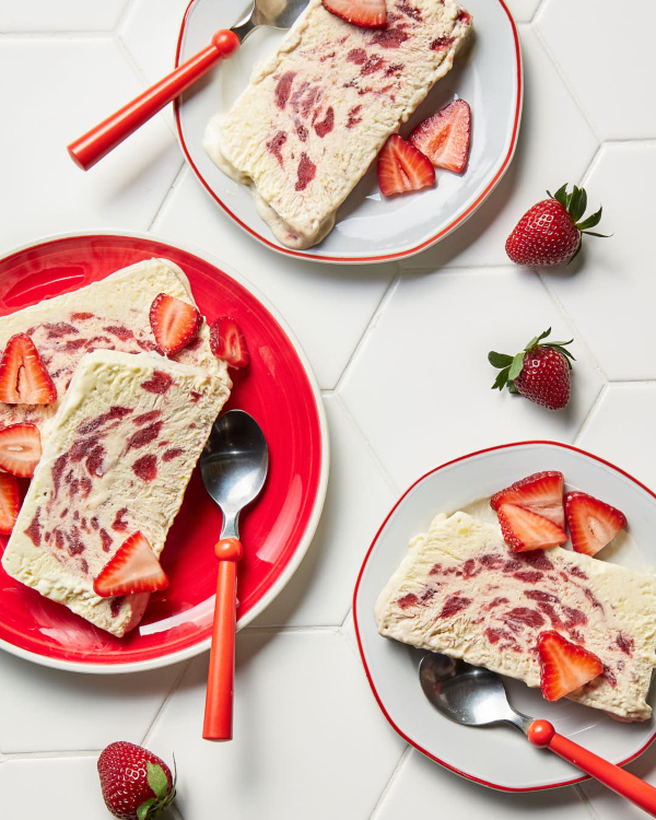  Des tranches de semifreddo à la fraise et à la fleur de sureau sur trois assiettes, deux blanches avec une bordure rouge et une rouge avec une bordure blanche, chacune avec une cuillère rouge, et présentées avec des fraises entières dispersées sur un comptoir en carrelage blanc