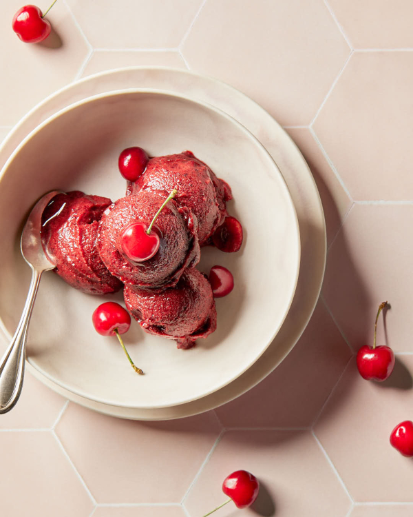 Quatre boules de sorbet aux cerises garnies de cerises, présentées sur un comptoir en carreaux hexagonaux rose pâle.