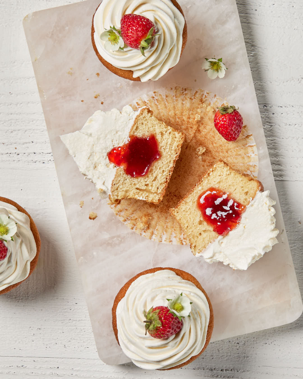 Quatre cupcakes à la vanille garnis d'un glaçage tourbillonnant et d'une fraise entière, l'un d'entre eux montré coupé en deux avec de la sauce aux fraises au centre, sur une planche à découper en granit.