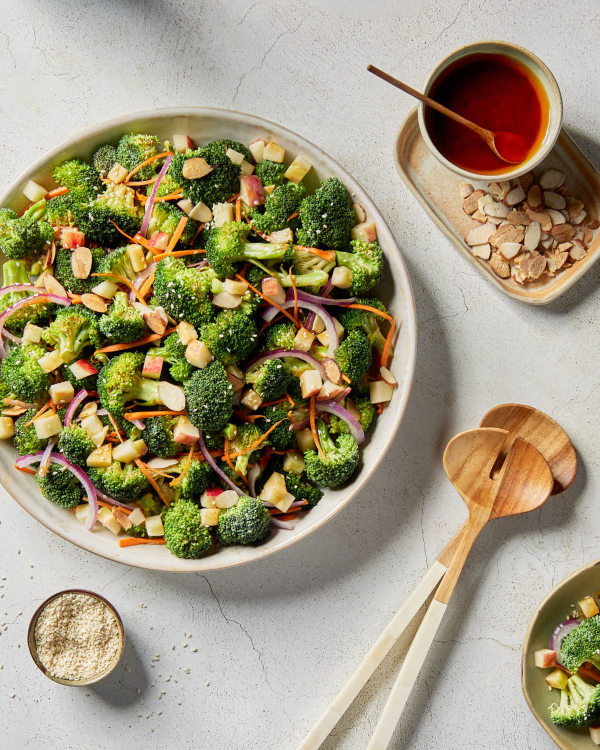 Un saladier de salade de brocoli avec une vinaigrette au gochujang est présenté sur un comptoir de cuisine, avec une carafe de vinaigrette et des cuillères de service en bois.