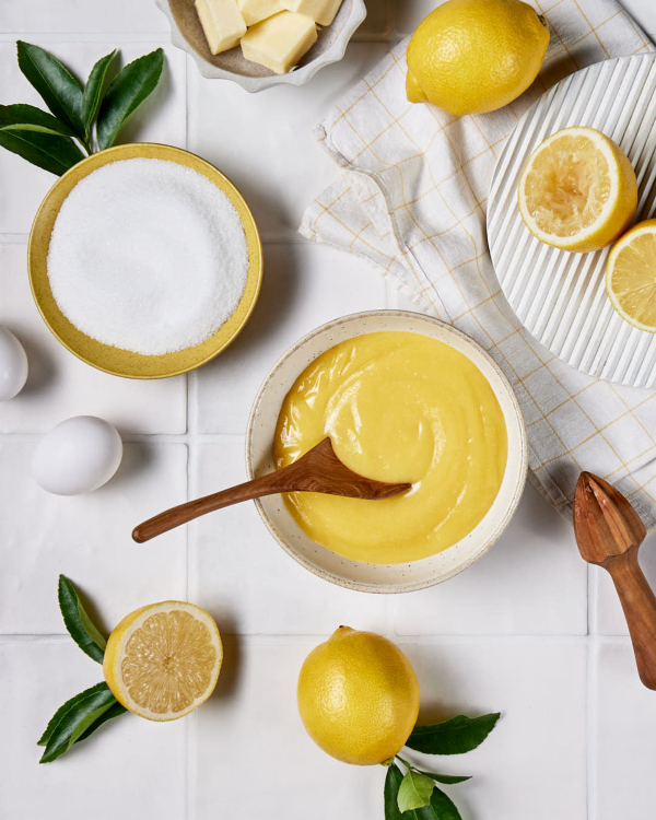 Un bol de lemon curd est présenté sur un comptoir carrelé, avec des citrons entiers et coupés en deux, un presse-citron et un bol de sucre granulé.