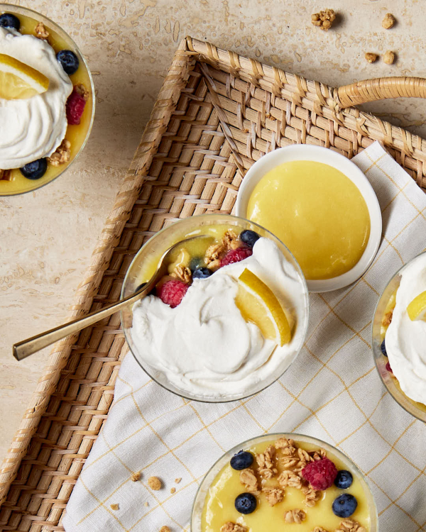 Un plat en verre de parfait au citron est présenté sur un plateau en osier avec un bol de lemon curd et trois autres parfaits.