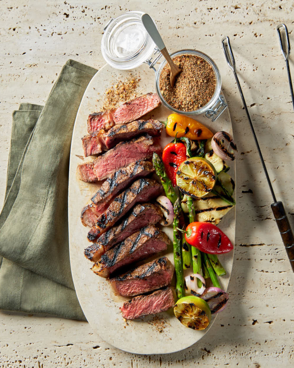 Un plateau de steak grillé et tranché ainsi que de légumes grillés, avec un bocal en verre avec un couvercle rempli d'épices sucrées pour steak à la montréalaise est présenté avec des pinces de barbecue sur un comptoir en calcaire.