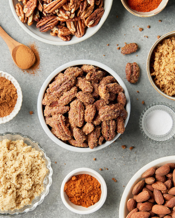 Un bol blanc débordant de pacanes et d’amandes confites, déposé sur un comptoir de cuisine avec une cuillère de bois et des bols d’amandes et de pacanes nature, de cassonade, de cannelle et de paprika.