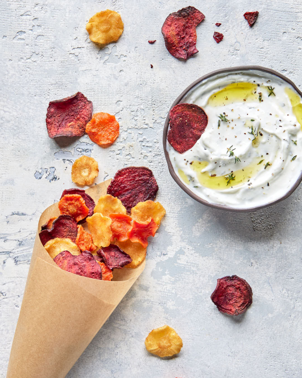 Un cône de papier parchemin rempli de croustilles de légumes racines mélangées avec plusieurs croustilles éparpillées autour et un bol de trempette crémeuse.