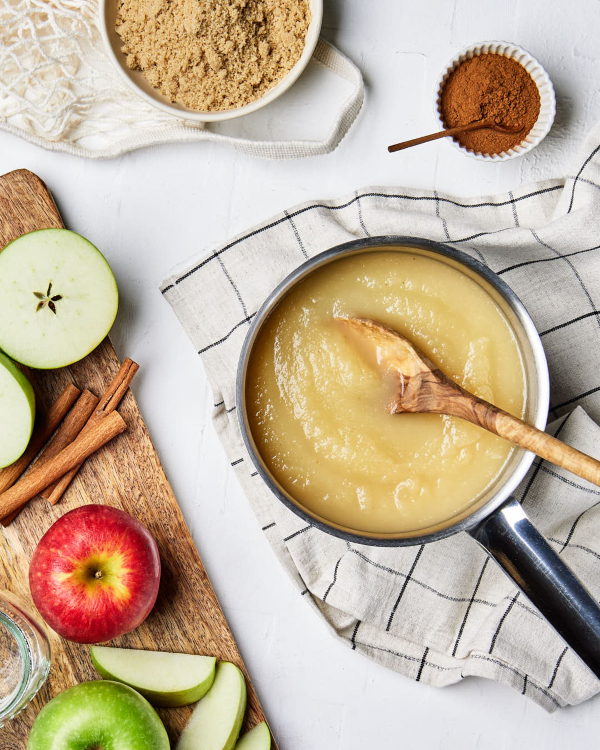 Une petite casserole de compote de pommes avec une cuillère de bois à l’intérieur, déposée sur un linge de cuisine avec des pommes entières et en tranches, des bâtonnets de cannelle, un bol de cannelle et un bol de Cassonade Dorée.