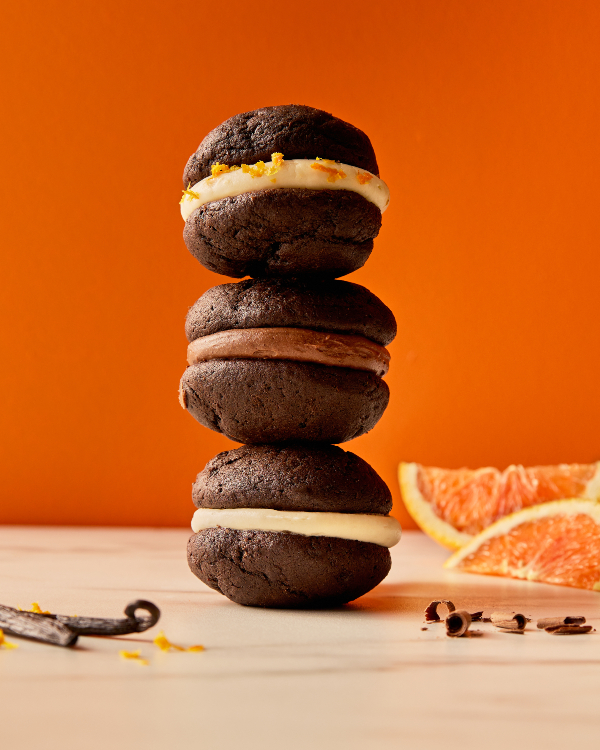 Trois mini whoopie pies au chocolat, chacun avec une garniture différente, empilés les uns sur les autres, présentés avec des gousses de vanille, du cacao râpé et des quartiers d'orange.