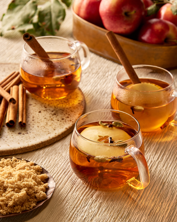 Trois tasses en verre de cidre de pomme chaud garnies de tranches de pomme, de clous de girofle et de bâtonnets de cannelle, présentées sur une table avec une carafe de cidre, un bol de pommes, et un bol de sucre jaune doré.