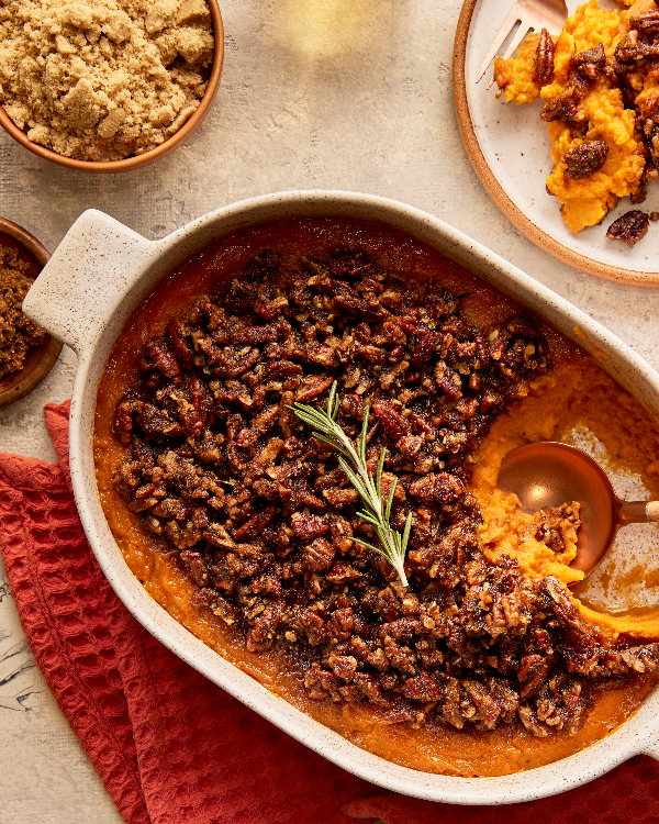 Une femme portant un chandail sert une casserole de patates douces garnie d'une branche de romarin depuis un plat de cuisson, présentée avec une salade de radicchio et d'agrumes, des verres de vin et des bols de sucre jaune doré et de sucre Demerara