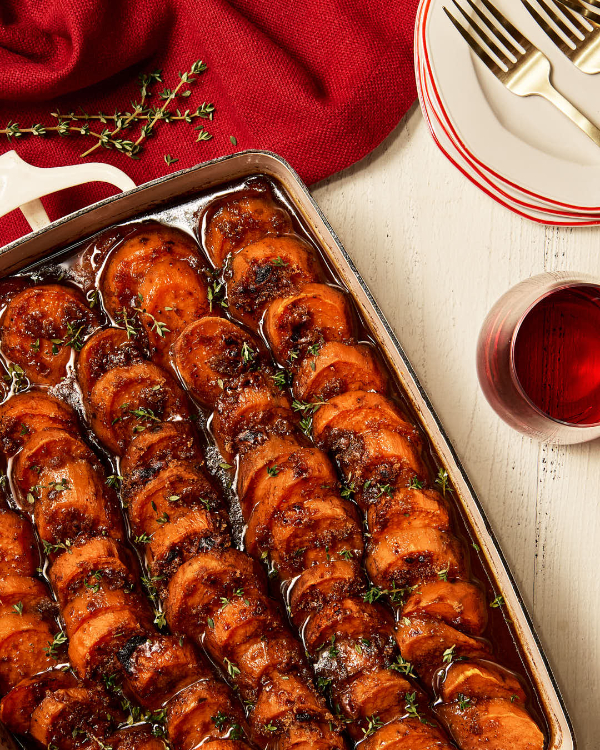 Des patates douces confites tranchées dans un plat à gratin garnies d'herbes et avec une caramélisation visible, présentées avec des assiettes, des fourchettes, et une boisson sombre dans un verre. 