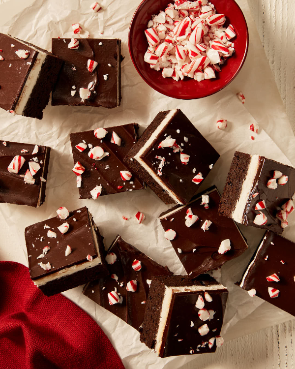 Des brownies au chocolat menthe avec une couche de crème à la menthe et un nappage de ganache, chacun saupoudré de morceaux de canne en bonbon, présentés sur du papier ciré sur une surface blanche, accompagnés d'une serviette rouge et d'un bol rouge de morceaux de canne en bonbon.