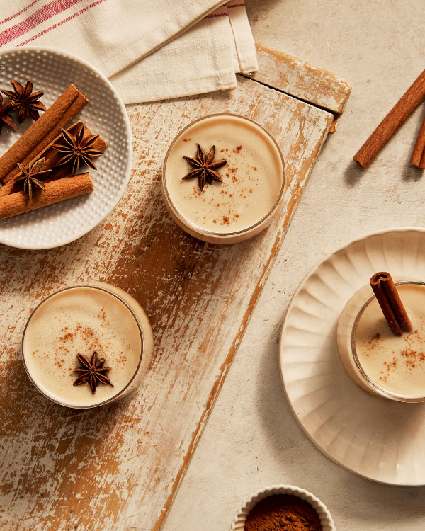  Deux verres tumblers de lait de poule sans œufs garnis d'anis et de cannelle, posés sur une planche à découper rustique en bois avec un troisième verre sur une soucoupe, présentés avec de la cannelle et de l'anis dans un plat.