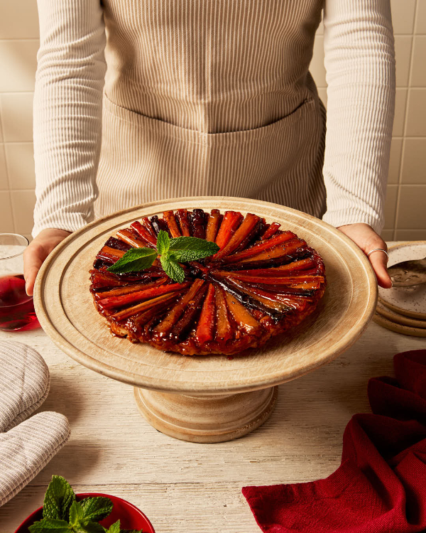 Une femme se tenant derrière une tarte tatin aux carottes et aux panais sur un présentoir à gâteaux, accompagnée de feuilles de menthe fraîche, de maniques, d'une boisson sombre et d'assiettes de service.