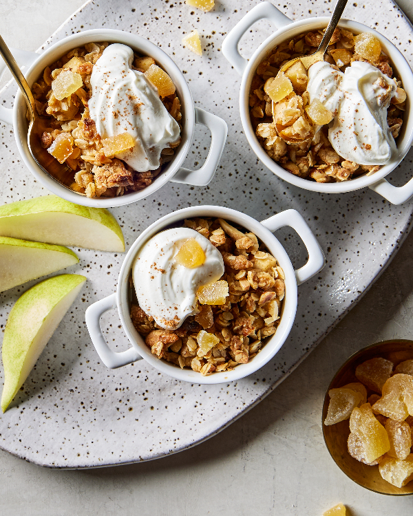 Vue aérienne de trois bols individuels blancs de crumble poire-gingembre avec crème fouettée, deux avec des cuillères dorées, présentés avec des tranches de poire et un petit plat de morceaux de gingembre.