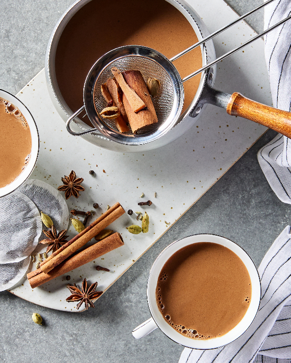 Vue de dessus de deux tasses de thé de masala chai et d'une théière de chai avec une passoire pleine d'épices entières, présentées avec des sachets de thé et diverses épices chaudes sur une planche à découper.