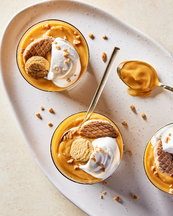 Vue de dessus de trois verres de pudding au caramel écossais sur un petit plateau ovale, chacun garni de crème fouettée, de morceaux de toffee et de biscuits.