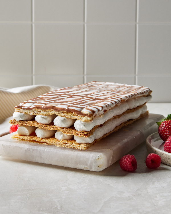 Un gâteau Mille-Feuille sur un plateau en marbre dans une cuisine avec un dosseret en carrelage, présenté avec des fraises et des framboises éparpillées.