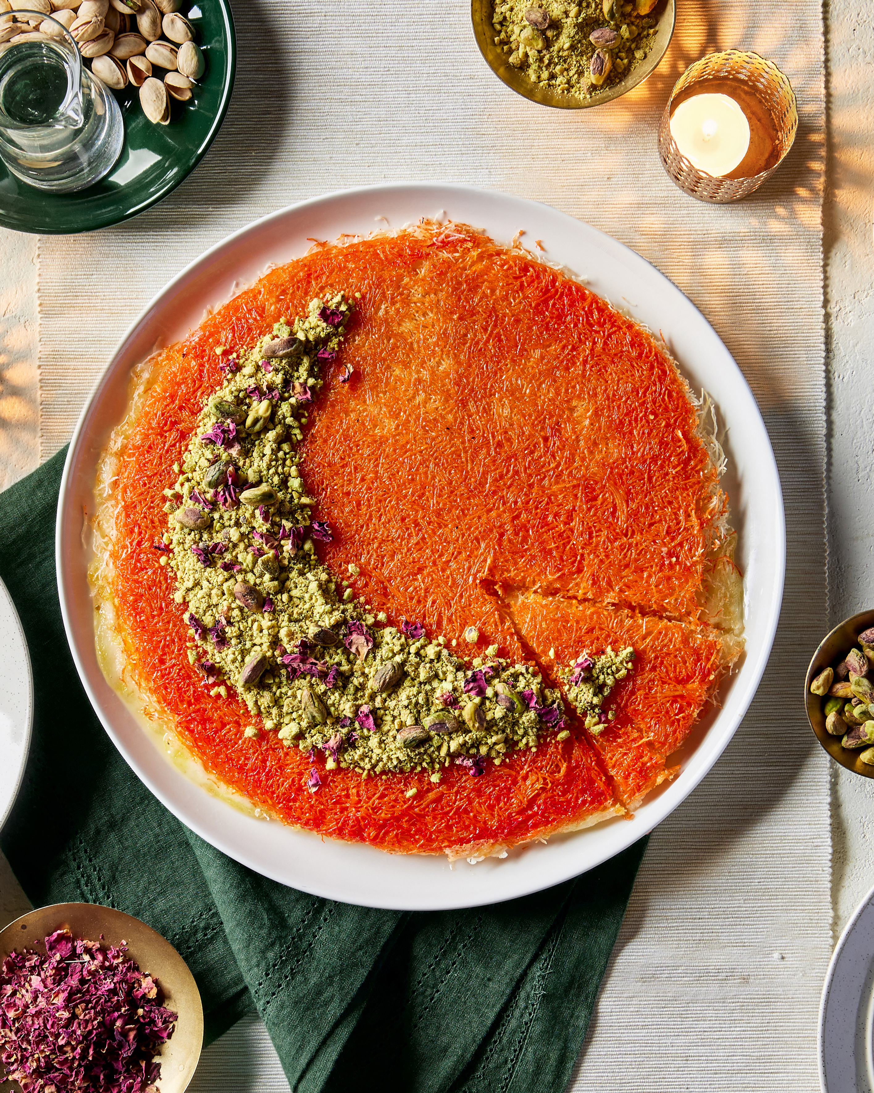 Knafeh dans une assiette avec une tranche coupée mais non retirée, garni de pistaches et de pétales de fleurs disposés en forme de croissant de lune, présenté avec des plats de pistaches en coque et de pétales de fleurs, et une bougie allumée.