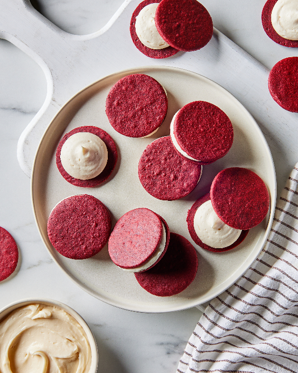 Un plat de biscuits sandwich velours rouge avec garniture à la crème sur un comptoir en marbre, à côté d'une serviette de cuisine rayée et d'un bol de garniture à la crème supplémentaire.