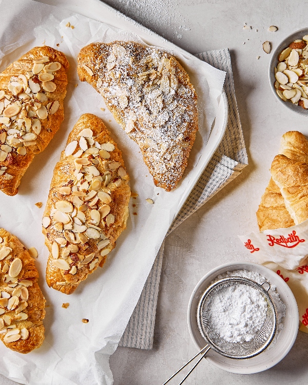 Vue de dessus de trois croissants aux amandes cuits à la friteuse à air avec un généreux nappage d'amandes tranchées et de sucre en poudre sur du papier parchemin, avec un croissant nature supplémentaire et un tamis plein de sucre en poudre sur une surface en marbre.