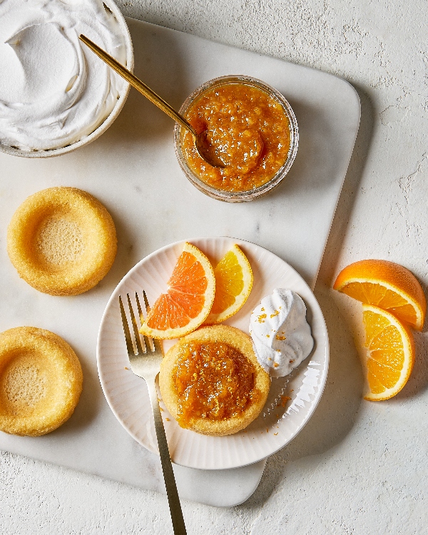 Vue de haut d'un cadre de dessert mettant en vedette un gâteau éponge avec un nappage de marmelade d'orange et un accompagnement de crème fouettée, des tranches d'orange fraîches sur l'assiette, des gâteaux éponge supplémentaires, et un bol de marmelade avec une cuillère en or, sur un comptoir en marbre.