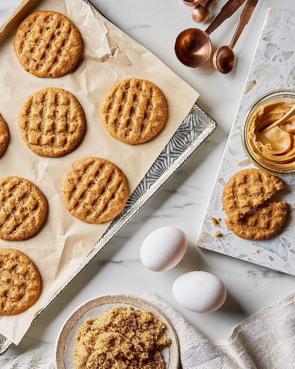 Huit biscuits au beurre d'arachide faits maison avec des surfaces dorées et des marques traditionnelles à la fourchette, accompagnés d'un pot de beurre d'arachide, de cuillères à mesurer et de deux œufs entiers sur un plan de travail de cuisine.