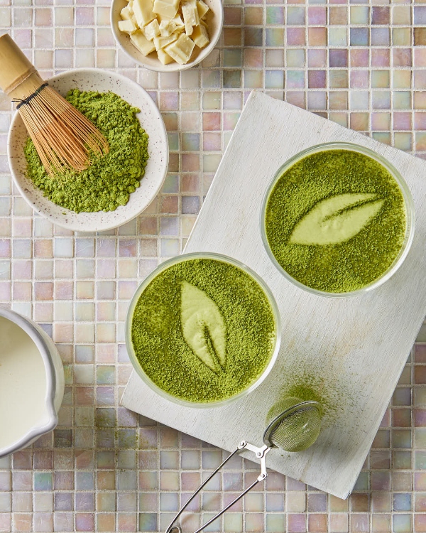 Vue aérienne de deux bols de mousse au chocolat blanc et matcha surmontés d'une fine couche de poudre de matcha, accompagnés d'ingrédients comme des morceaux de chocolat blanc et un pichet de crème, avec un fouet en bambou et une passoire sur une planche en bois blanc, le tout disposé sur une surface carrelée multicolore.