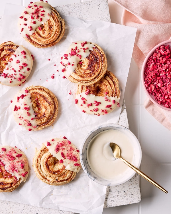 Biscuits à la pâte feuilletée aux framboises glaçés au chocolat blanc et saupoudrés de framboises lyophilisées, un bol de framboises écrasées, un bol d’enrobage au chocolat blanc et une cuillère dorée sur papier parchemin montré sur une planche à découper en marbre sur un comptoir de tuiles blanches.