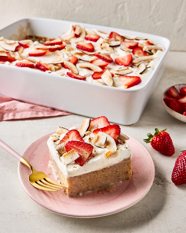 Une part de gâteau tres leches aux fraises et à la noix de coco sur une assiette rose avec une fourchette dorée, à côté d’un moule blanc en céramique contenant le même gâteau et d’un petit bol de fraises tranchées, le tout sur une surface texturée grise avec une serviette en lin rose.