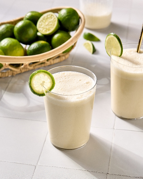  Deux verres de limade brésilienne crémeuse, garnis chacun d'un quartier de lime fraîche. Un des verres contient une paille. Les breuvages reposent sur une surface de carreaux pâles, avec un panier en osier rempli de limes entières et de moitiés de lime en arrière-plan. On voit aussi d'autres quartiers de lime et un pichet de limade.