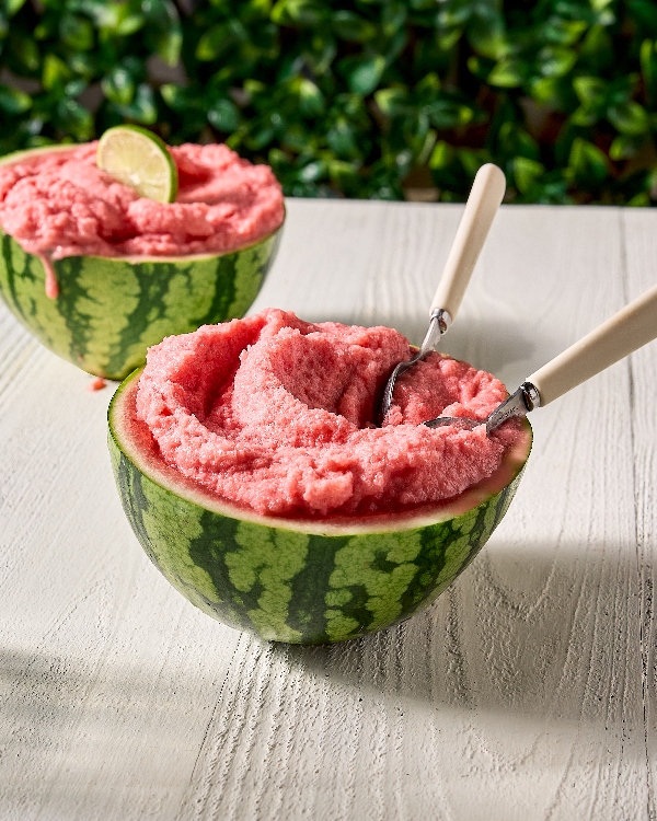 Deux moitiés de melon d'eau évidées, remplies de sorbet à la pastèque rose et onctueux, reposent sur une table en bois blanc. Deux cuillères à melon en inox se trouvent posées dans la moitié avant du melon. La moitié arrière du melon est décorée d'un quartier de lime. Le tout est mis en valeur par un arrière-plan de feuillage verdoyant.