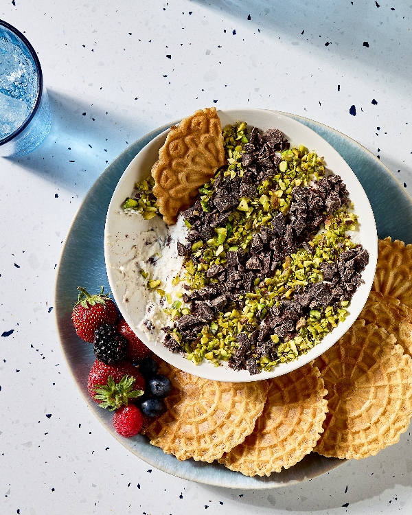 Vue plongeante sur une assiette posée sur un comptoir en calcaire. L'assiette présente un bol de trempette à cannoli garni de pistaches hachées et de morceaux de chocolat. Des biscuits Pizzelle et des fruits frais - des fraises, des mûres, des framboises et des bleuets - sont disposés autour du bol. Un verre d'eau pétillante est visible sur le côté gauche.