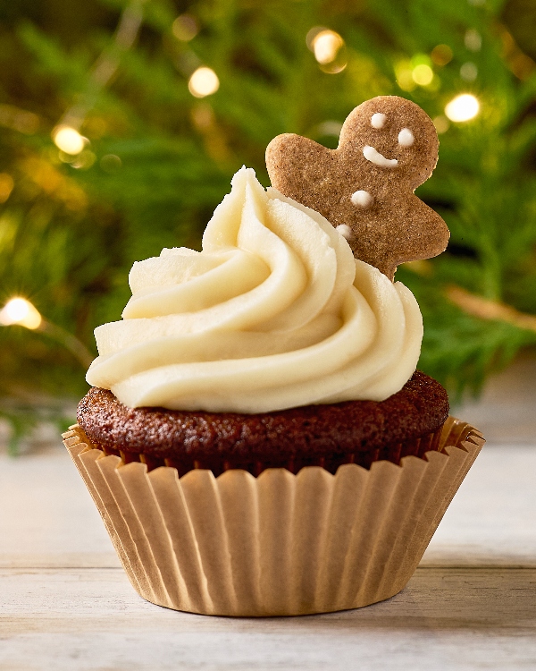 Gros plan sur un cupcake au pain d'épices avec un tourbillon de glaçage blanc, surmonté d'un mini biscuit au pain d'épices. L'arrière-plan présente une guirlande verte festive avec des lumières féériques douces et chaudes, mettant en valeur le thème des fêtes.