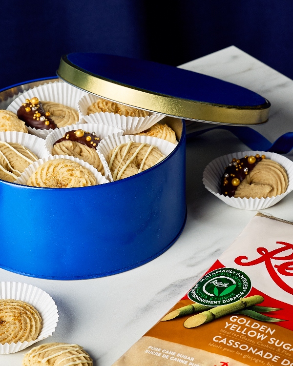 Une boîte-cadeau remplie de biscuits au beurre décorés et disposés dans des moules en papier blanc. Les biscuits présentent une variété de décorations, notamment trempés dans du chocolat avec des paillettes dorées, nappés de chocolat blanc et saupoudrés de sucre doré. Un sac de sucre jaune doré est partiellement visible sur un comptoir en marbre, rehaussant le thème de la pâtisserie.