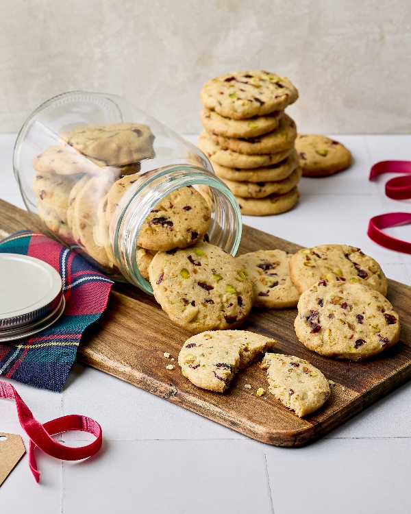 Des biscuits à la canneberge et à la pistache débordant d'un pot de verre sur une planche de bois. Une pile de biscuits et un sac d'ingrédients de cuisson sont visibles en arrière-plan. Une serviette de table à carreaux rouges et verts, ainsi qu'un ruban et une étiquette-cadeau, suggèrent un thème de cadeaux de Noël. Un biscuit est cassé en deux, révélant un intérieur moelleux avec des morceaux de canneberges et de pistaches