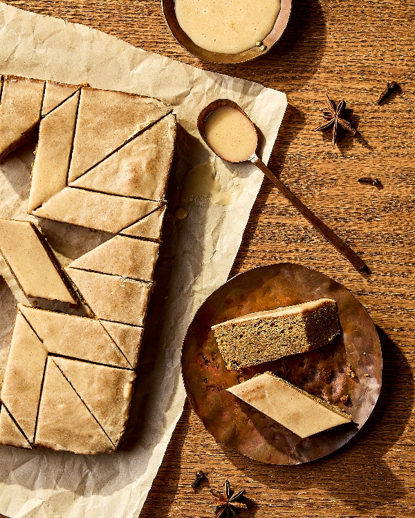 Des barres de gâteau épicées disposées sur du papier parchemin, coupées en losanges nets et recouvertes d'un glaçage brillant. Une assiette en métal contient une seule barre coupée en deux, mettant en valeur la mie tendre du gâteau. Une cuillère en bois avec du glaçage supplémentaire, un petit bol et des épices entières comme l'anis étoilé et les clous de girofle sont disposés à proximité, soulignant les saveurs chaudes et épicées du dessert