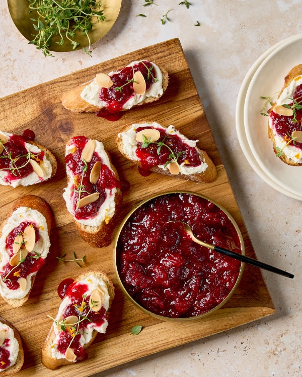 Une planche de bois remplie de crostinis garnis de feta fouettée, de chutney de canneberges, d'amandes effilées et de thym frais. Un petit bol de chutney de canneberges se trouve également sur la planche.