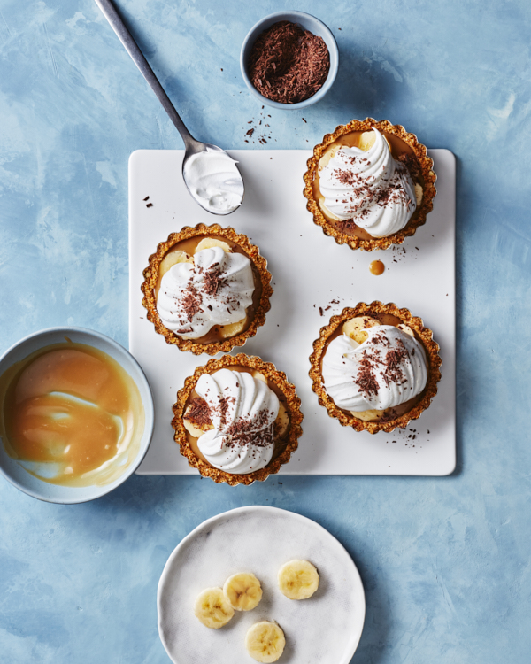 Quatre tartes banoffee sur un plateau blanc, accompagnées d'un bol de garniture au caramel, d'un bol de chocolat râpé et de tranches de banane sur une assiette.