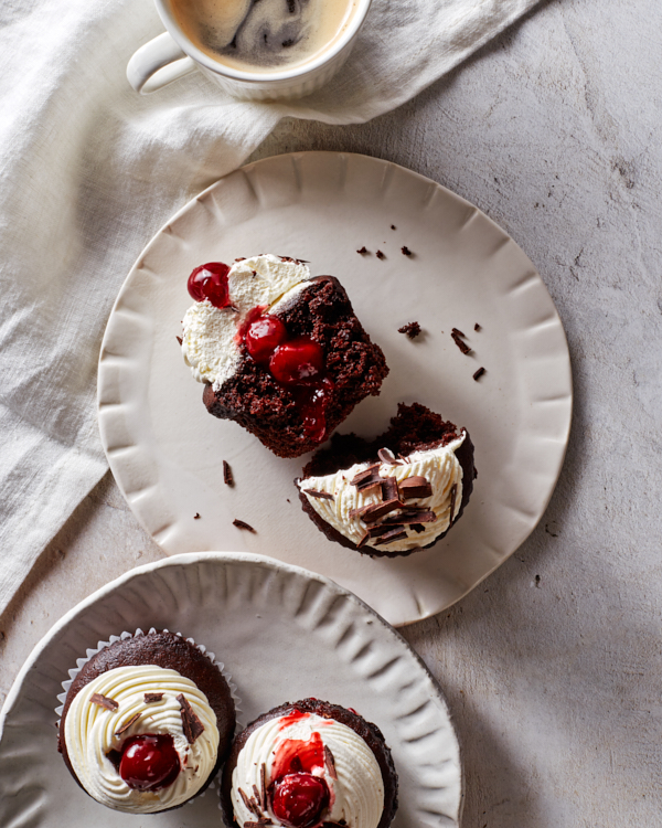  Trois petits gâteaux Forêt Noire avec glaçage à la chantilly, garnis de cerises et de chocolat râpé ; deux sur un plateau et un coupé en deux sur une assiette.