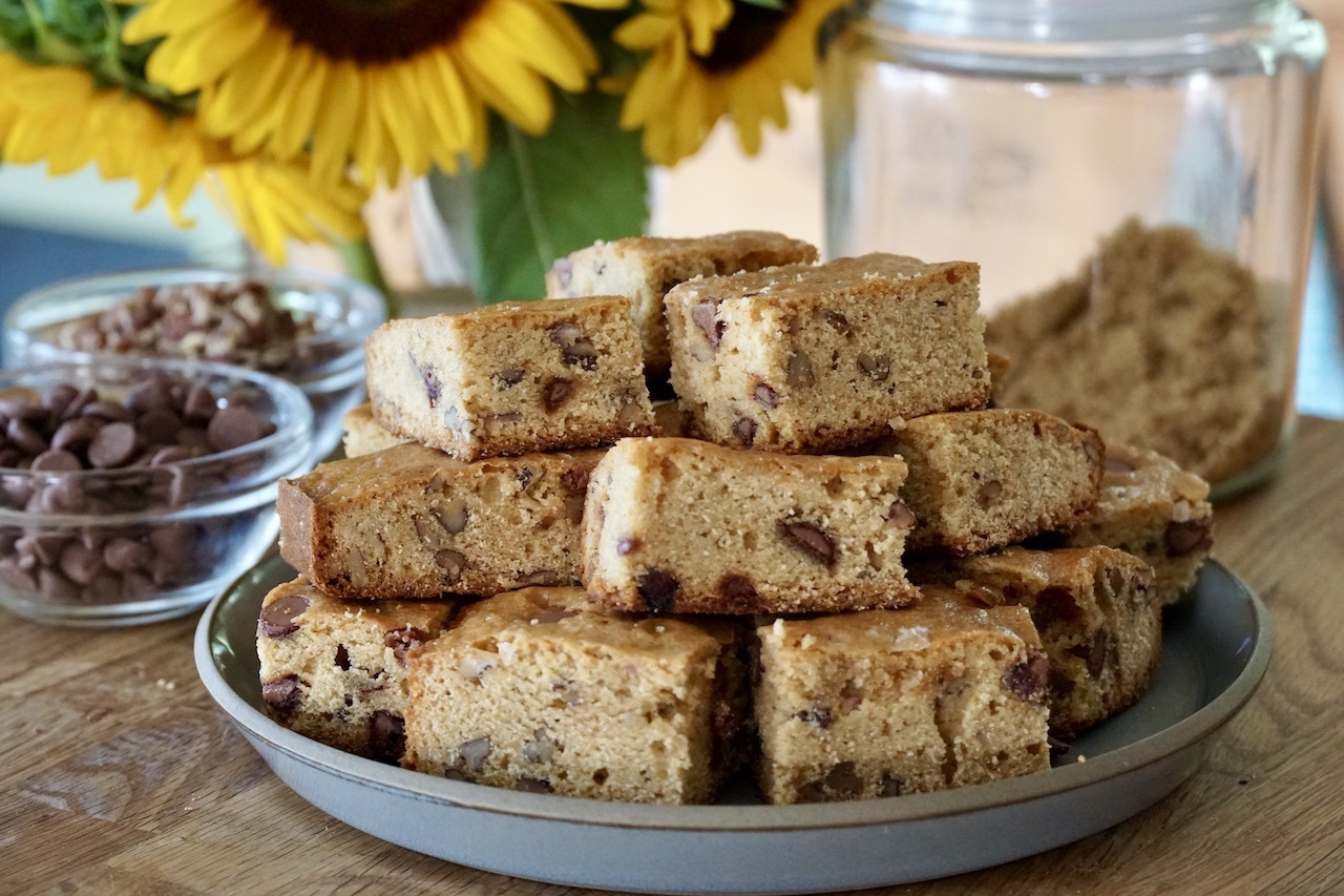 Blondies au beurre noisettes couronnés de pépites de chocolat disposés sur un comptoir de type bloc de boucher avec des tournesols