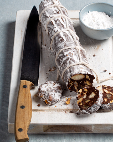 Salami au chocolat et aux noix saupoudré de sucre à glacer sur une planche à découper en bois avec un couteau, un bol de sucre à glacer et trois tranches coupées