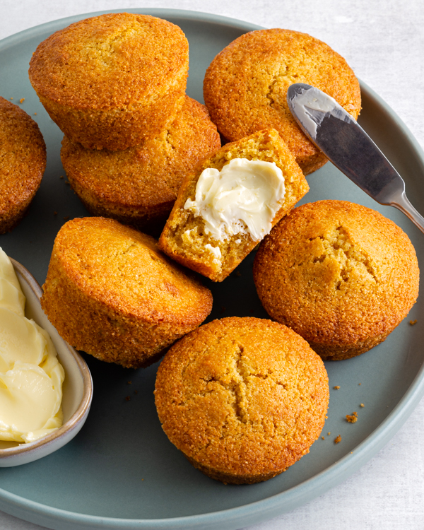 Des muffins à la semoule de maïs sur une assiette, dont un est coupé en deux et tartiné de beurre, avec un beurrier et un couteau