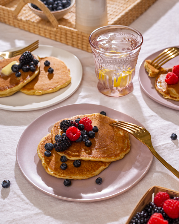 Plan en plongée de trois assiettes de pancakes garnis de petits fruits, disposées sur une table avec des ustensiles dorés, des petits fruits et du sirop en extra, et un verre d’eau citronnée.