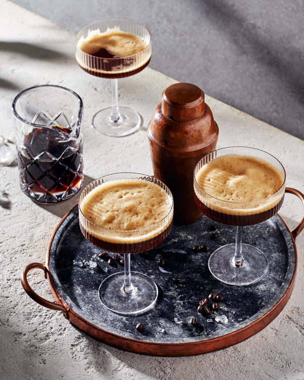 Deux verres de martini espresso mousseux sur un plateau avec un shaker à cocktail avec un troisième verre et un pichet en verre de martini espresso en arrière-plan.