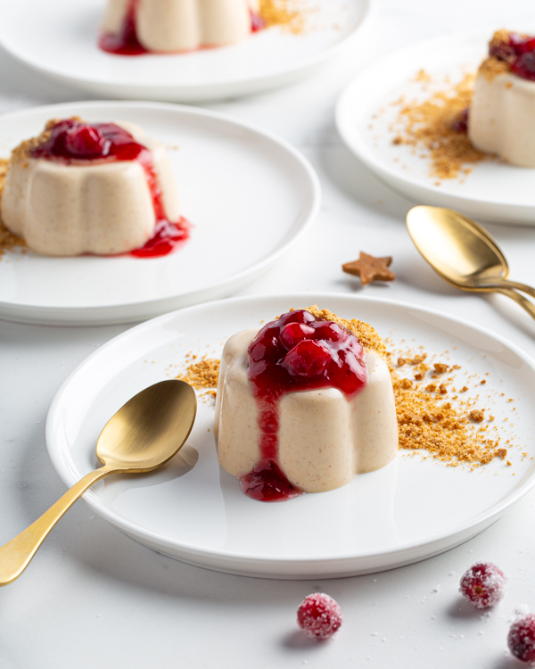 Quatre panna cottas au pain d’épices et babeurre sur des assiettes garnies de miettes de pain d’épices et de sauce canneberges au vin