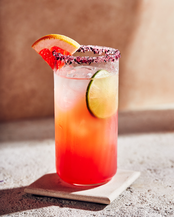 Un verre de paloma à l'hibiscus garni d'un quartier de pamplemousse et bordé de pétales d'hibiscus.